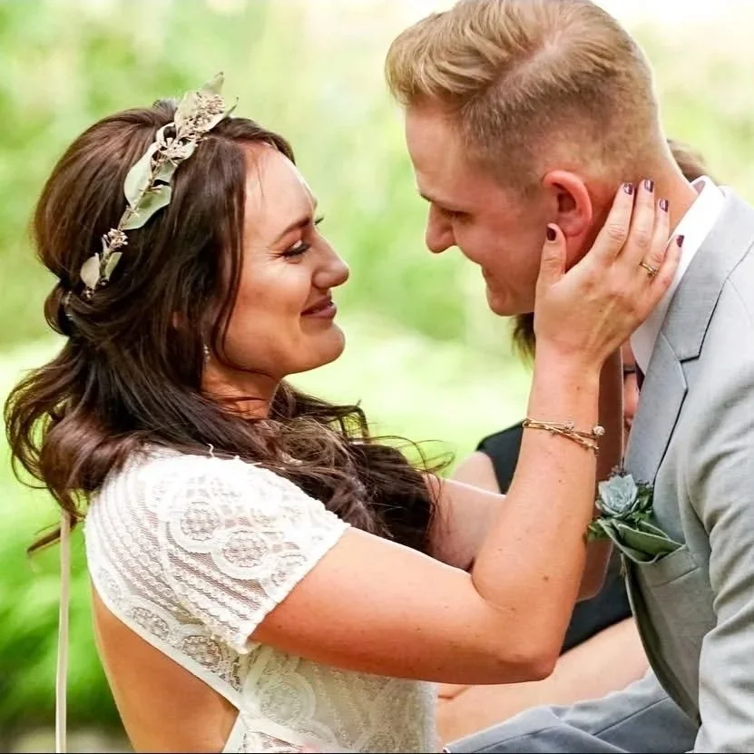 A woman and a man are facing each other in a close, intimate moment, possibly during a wedding ceremony. The woman is gently holding the man's face with both hands, and they are smiling lovingly at each other. The woman has long brown hair with a flo