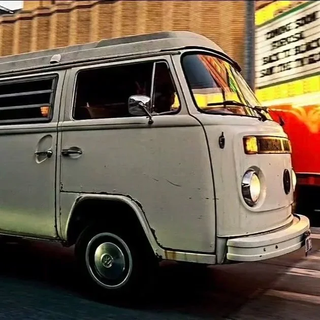 A vintage silver Volkswagen Type 2 van driving past a colorful billboard on a city street