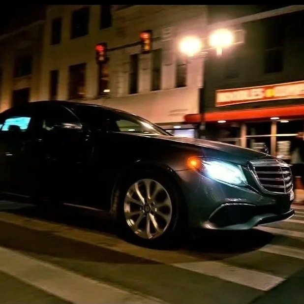 A black luxury sedan driving on a city street at night, with buildings and streetlights in the background.