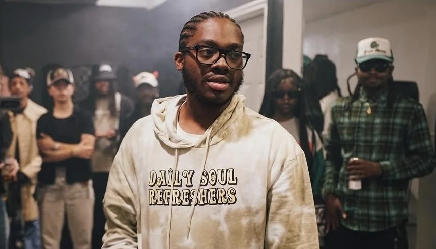 A man with glasses and braids in his hair stands in the foreground of a room filled with people, some of whom are wearing hats and casual clothing, at an indoor social or event setting.