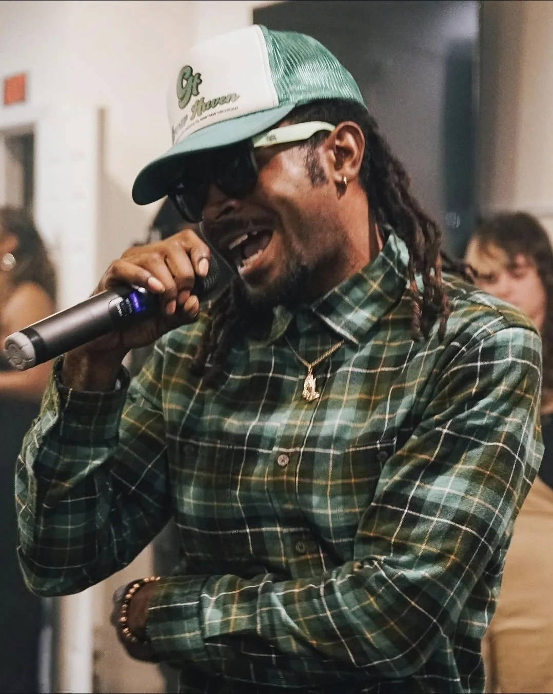 A man with dreadlocks, wearing a green and black plaid shirt, a white and green trucker hat, sunglasses, gold jewelry, and a microphone, singing or speaking passionately at an indoor event.