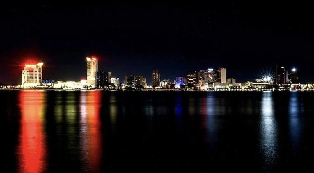Nighttime city skyline with illuminated buildings and reflections on water.