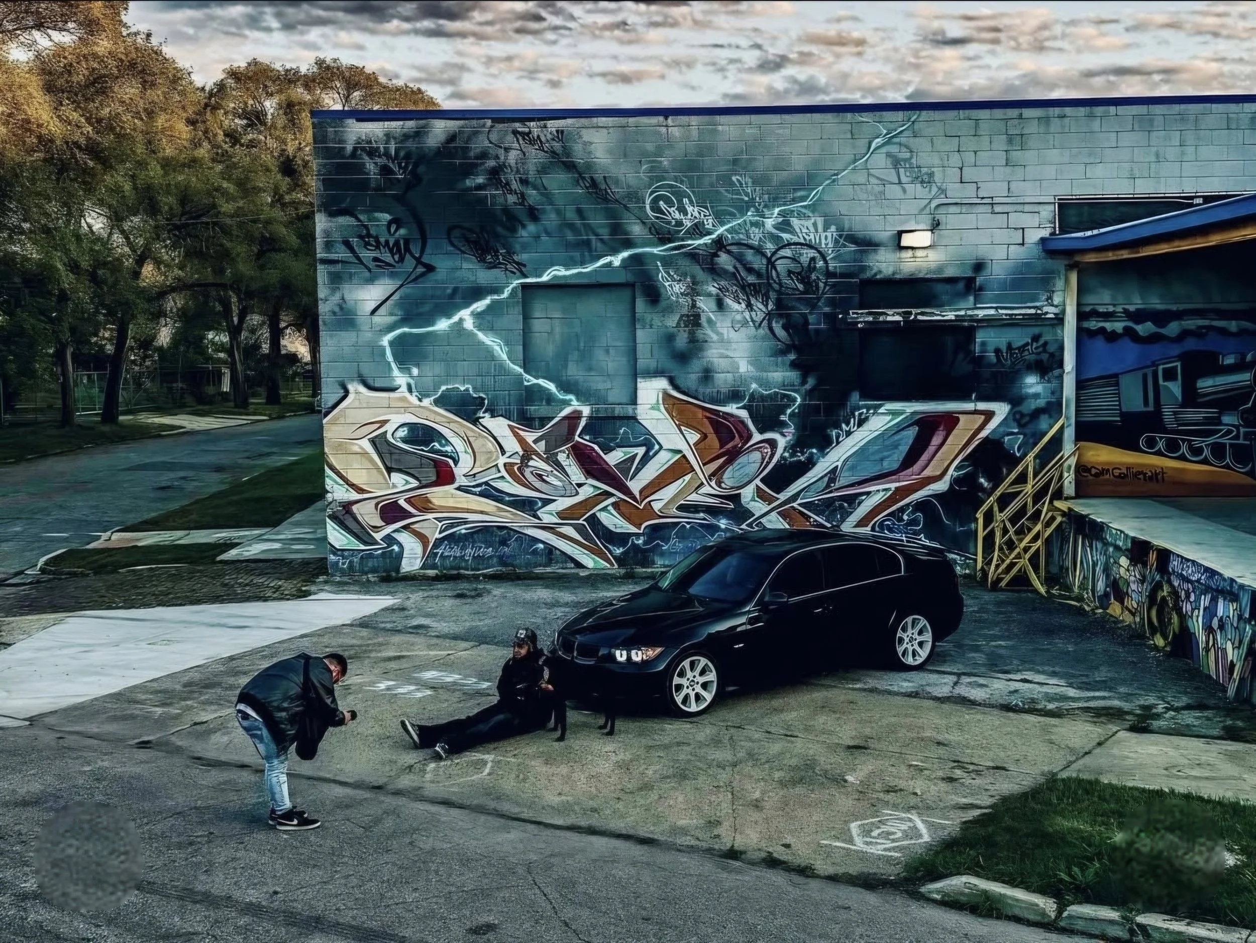 Two people, one taking a photo, in front of a black car parked on an urban scene with graffiti murals on the wall behind them.
