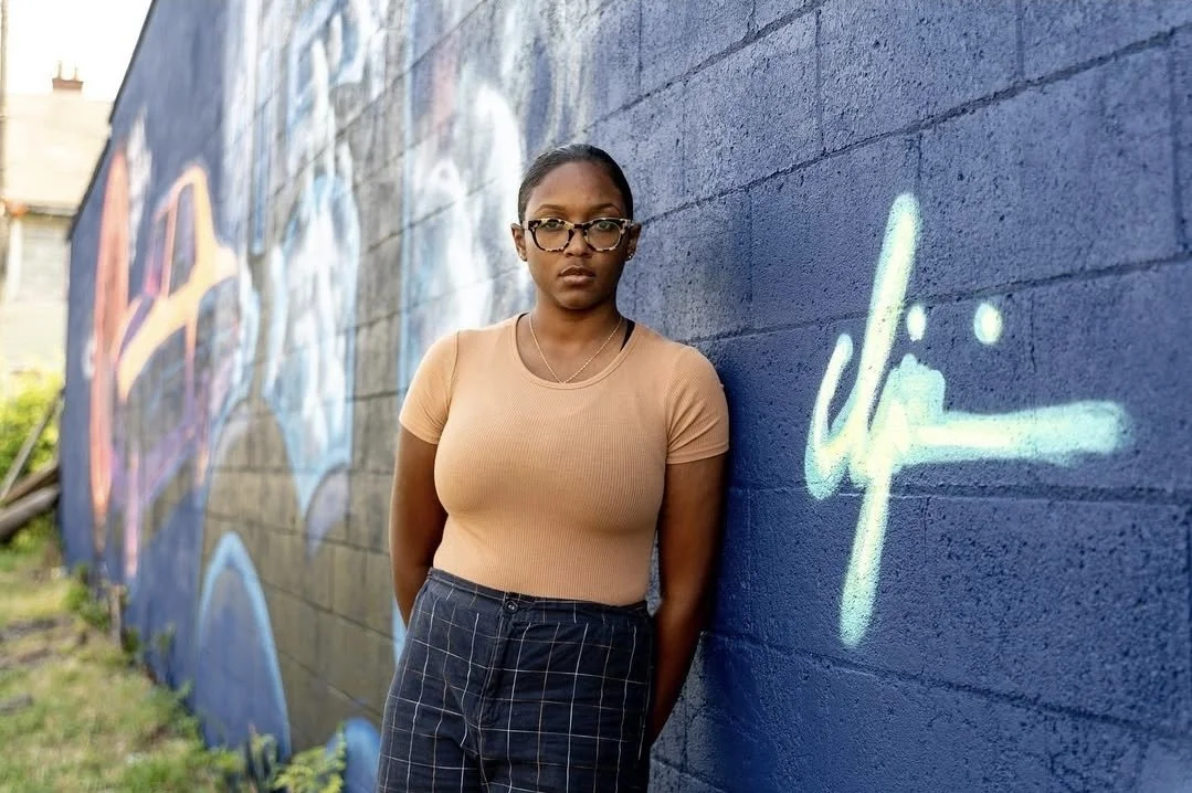 A woman with glasses, wearing a beige t-shirt and dark checkered pants, standing against a graffiti-covered blue brick wall.