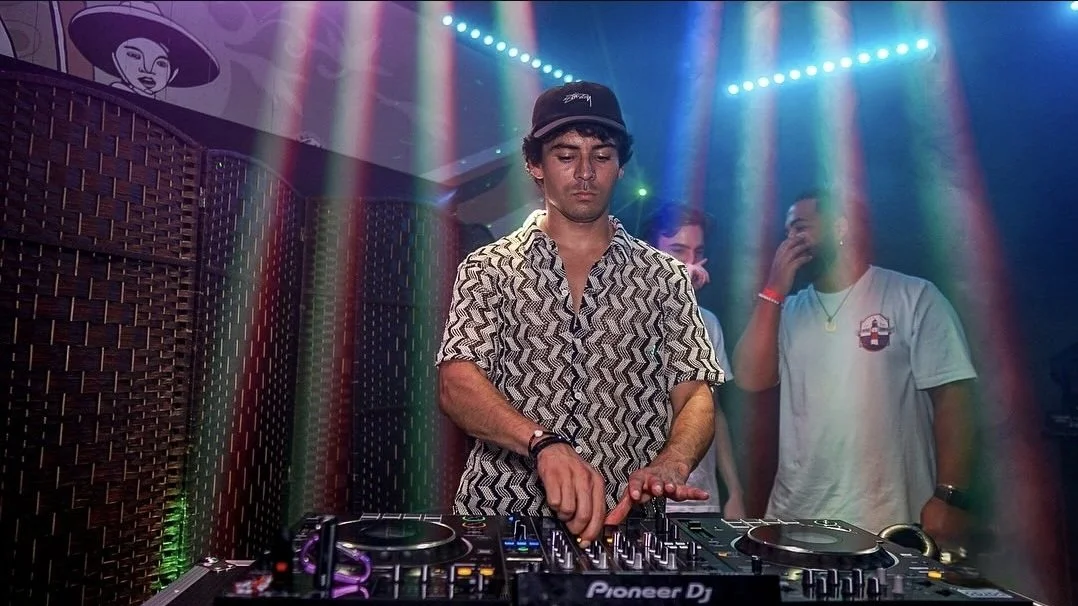 DJ wearing a patterned shirt and black cap performing at a nightclub with colorful lights and people in the background.