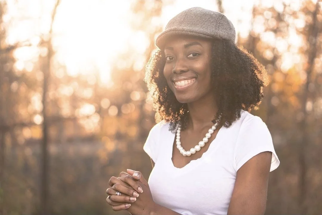 A smiling woman with curly hair, wearing a hat, pearl necklace, and white shirt, standing outdoors during sunset.