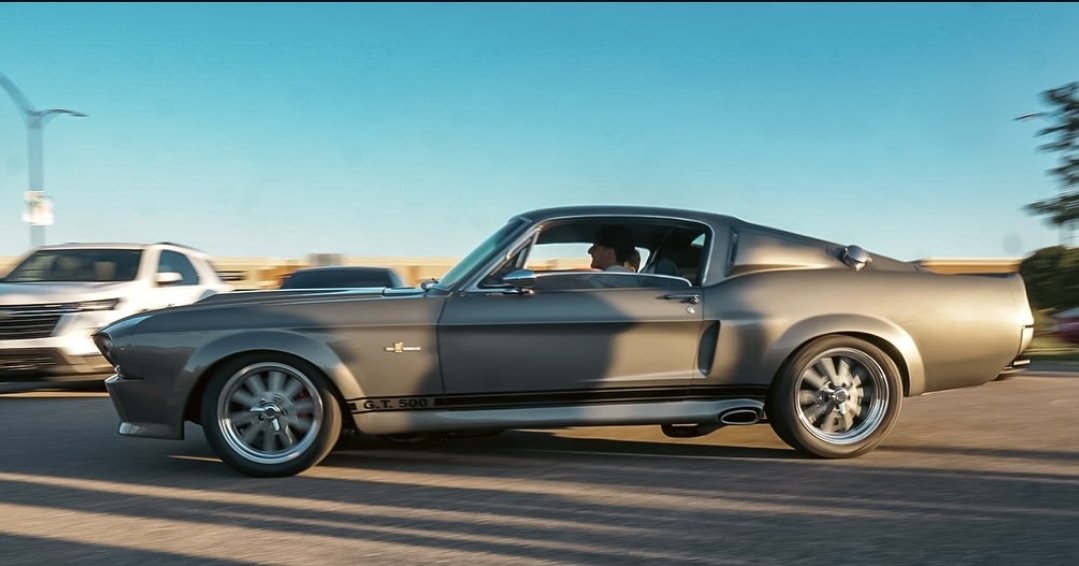 A classic black Ford Mustang G.T. 500 driving on a highway with a modern white SUV in the background.