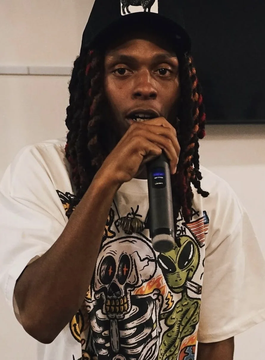 A young man with dreadlocks wearing a black cap and a colorful graphic t-shirt, holding a microphone and speaking.