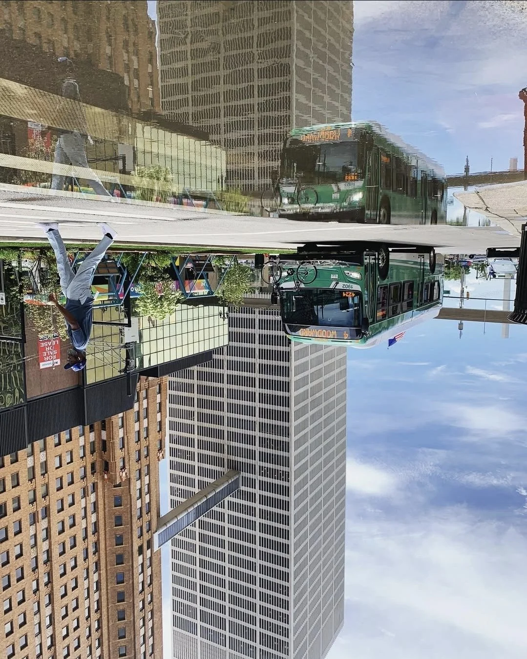 City street scene with a reflective glass building, a green bus, bicycles, and a man holding a phone, with skyscrapers and a blue sky in the background.