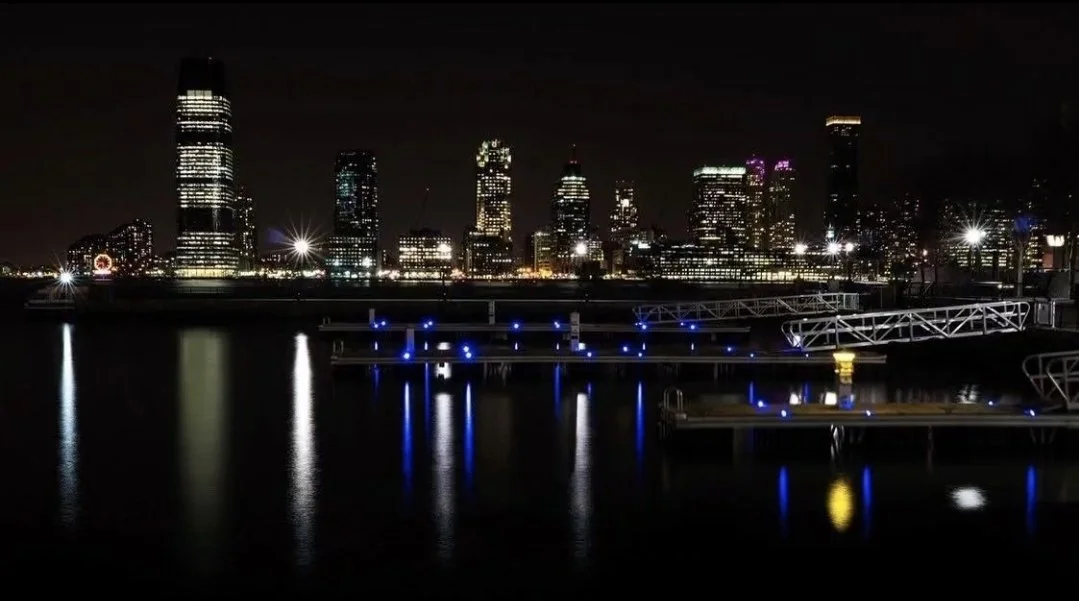 Nighttime cityscape with tall buildings lit up, reflecting on the water, with docks and blue lights in the foreground.