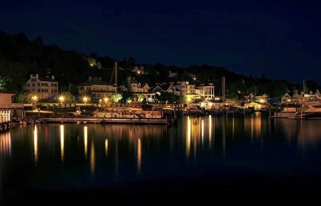 Nighttime view of a harbor with boats and yachts, illuminated houses, and their reflections on calm water.