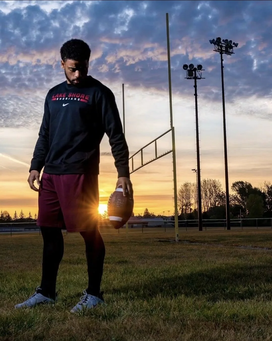 A young man standing on a football field at sunset, holding a football in his right hand, wearing a black long-sleeve shirt with 'LAKE SHORE SHORIANS' written on it, maroon shorts, black leggings, and white sneakers.