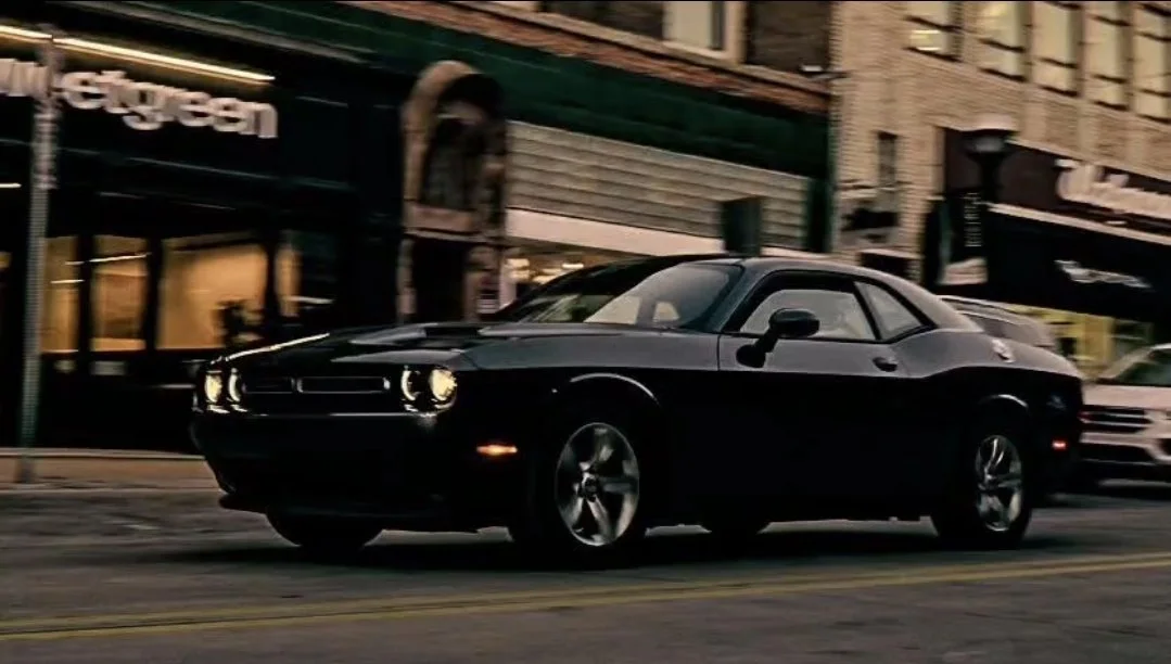 A black muscle car driving on a city street at night, with buildings and storefronts in the background.