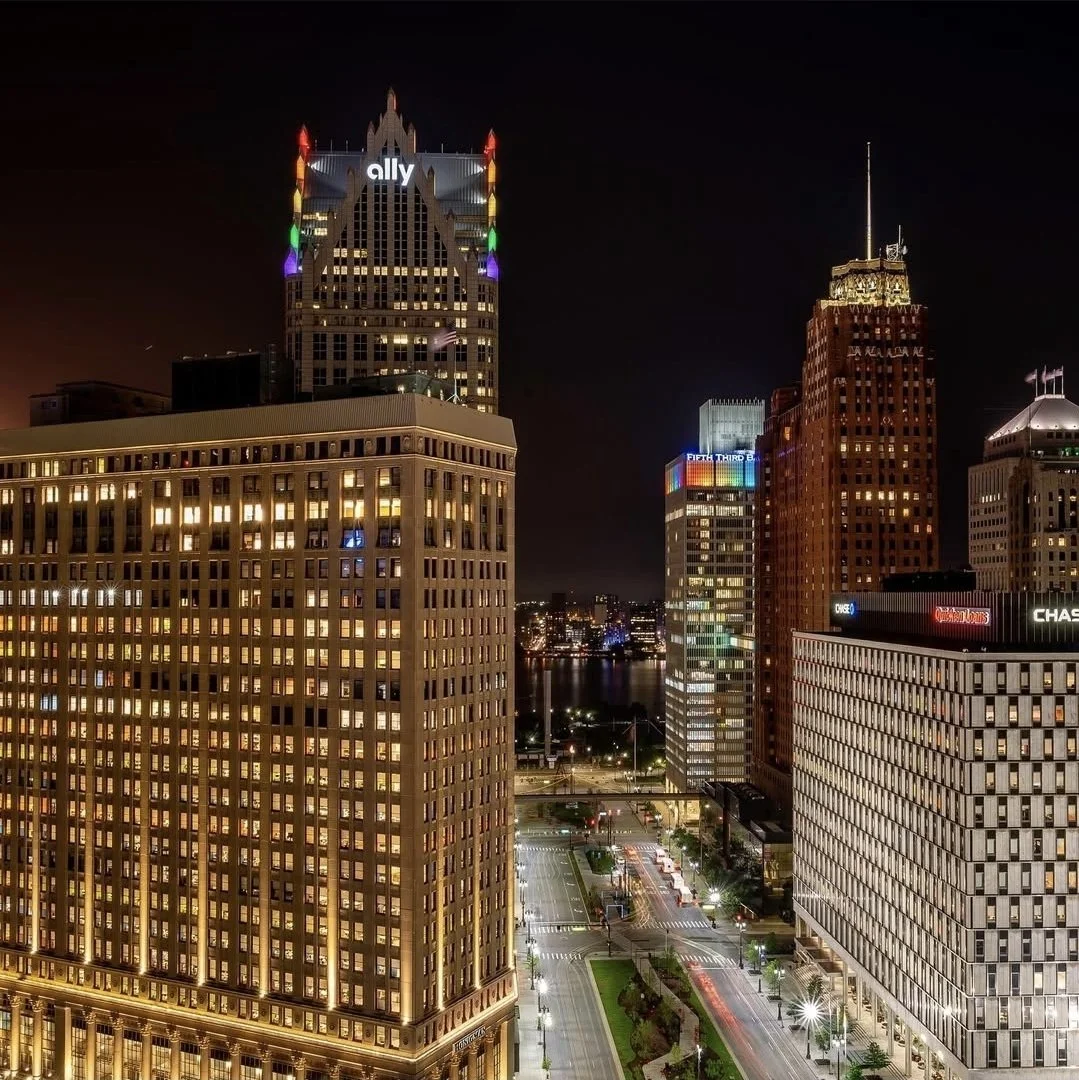 Nighttime cityscape of downtown with illuminated tall buildings, including one with colorful lights at the top and an 'ally' sign, and busy streets with moving lights and streetlights.