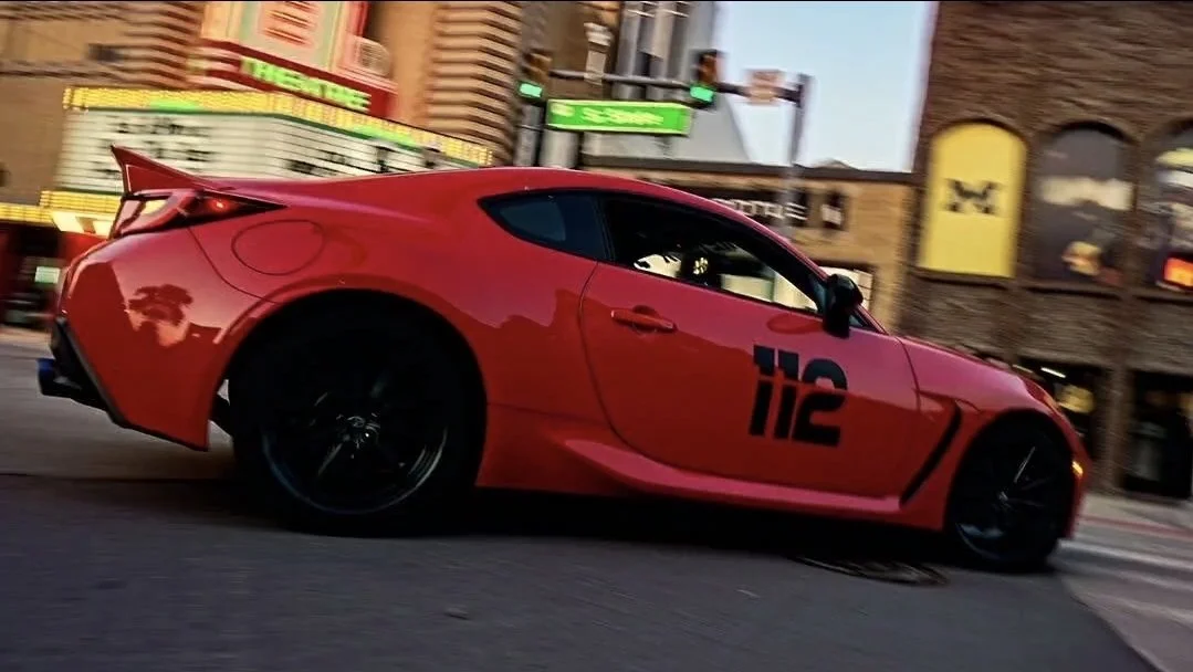 A red sports car driving on a city street at dusk with buildings and neon signs in the background.