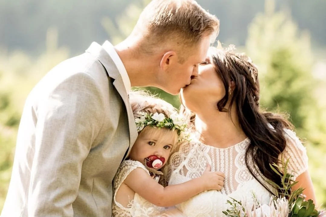 A couple in wedding attire sharing a kiss outdoors, with a young child dressed in white, wearing a floral headband and pacifier, in their arms.