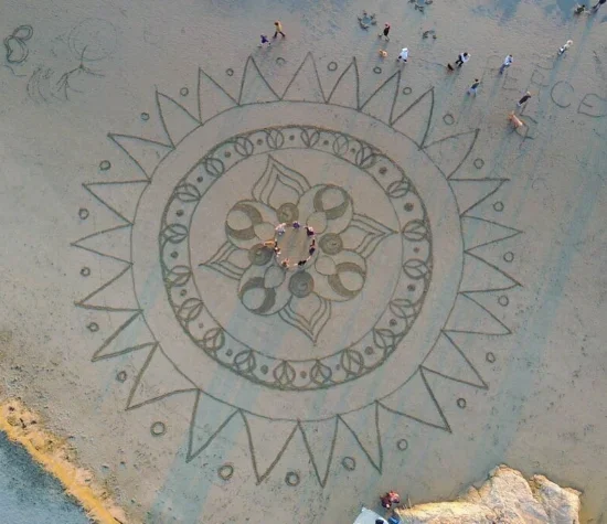 Beach with a large decorative sand art design featuring intricate patterns and a circle of people gathered in the center.