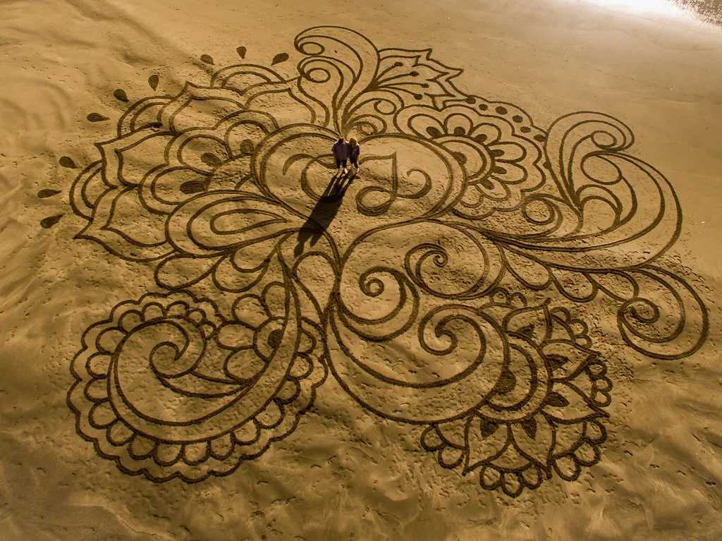 A large sand art design with intricate floral and swirl patterns, created on a sandy surface, with two people standing in the center.