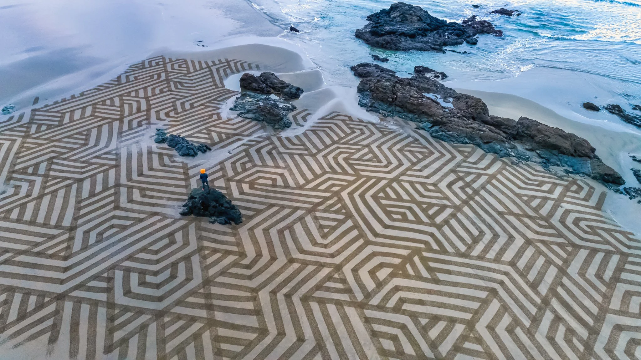 A person stands on a rocky outcrop overlooking an elaborate geometric pattern on a sandy beach, with rocks and the ocean in the background.