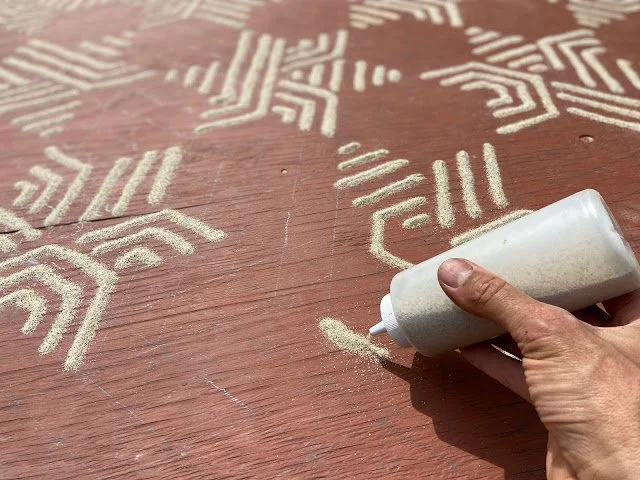 A person applying decorative white sand or powder through a tube onto a wooden surface to create geometric patterns or designs.