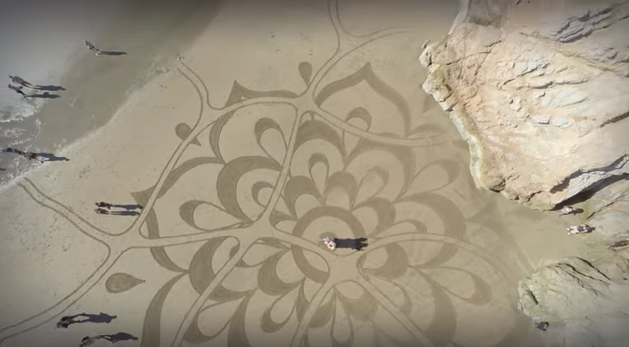 A large sand art pattern resembling a mandala on a beach with a few people and rocks around.