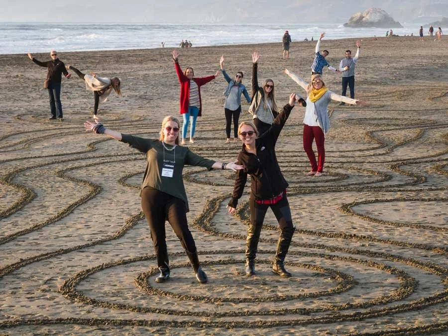 Group of people on a sandy beach with a maze pattern, waving and smiling at the camera, ocean and rocks in the background.