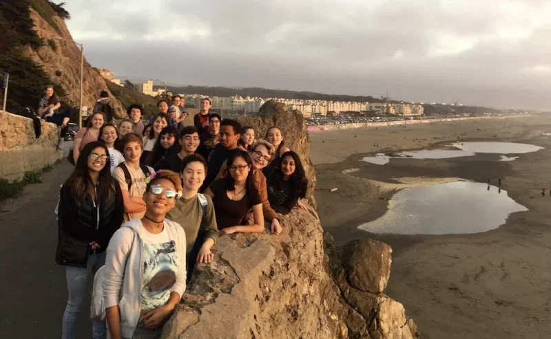 A group of people standing on a rocky cliff overlooking a beach and ocean during sunset.