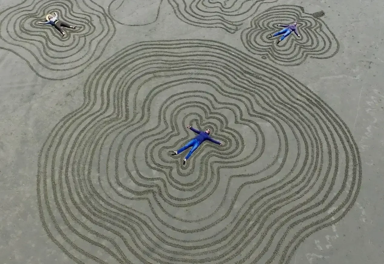 Three people lying on a large ground drawing of topographic contour lines, with the person in the center forming the middle of the lines and the other two positioned at the edges of the drawing.