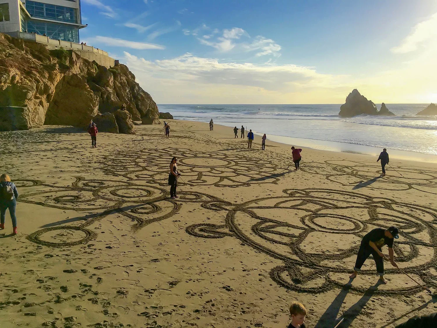 People drawing large intricate patterns in the sand on a beach with rocky cliffs and ocean waves in the background during sunset.