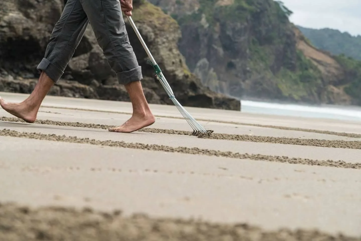 Person using a rake on a sandy beach, walking barefoot, with cliffs and ocean in the background.