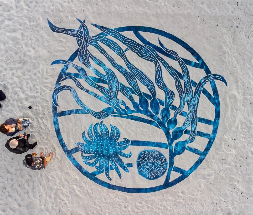 A large sand art of a circular design featuring a stylized tree with long, twisting branches and two abstract sea creatures or flowers at the base, created on a sandy surface with four people standing nearby.
