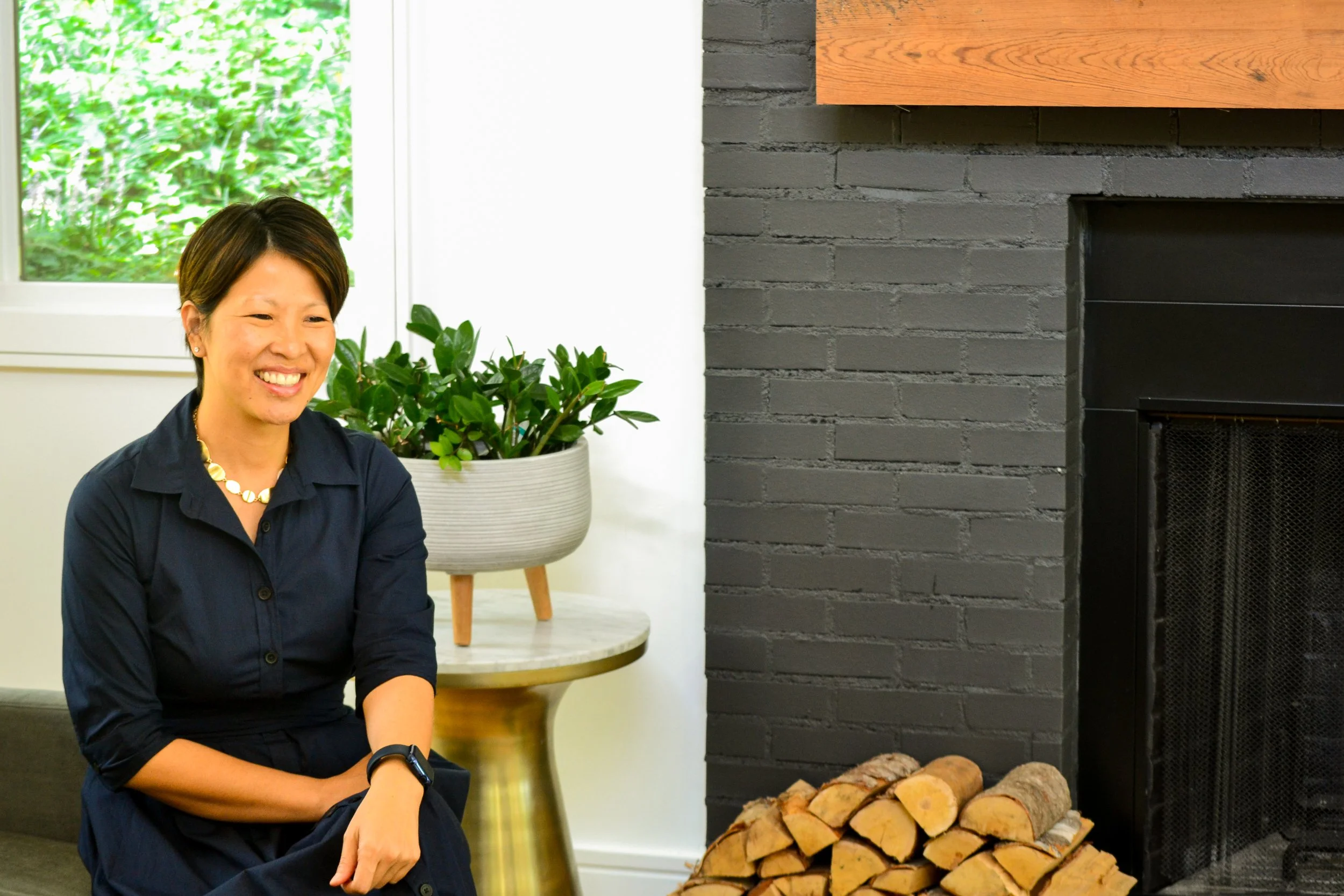A woman sitting indoors near a fireplace with stacked firewood, a potted plant on a side table, and a window showing greenery outside.