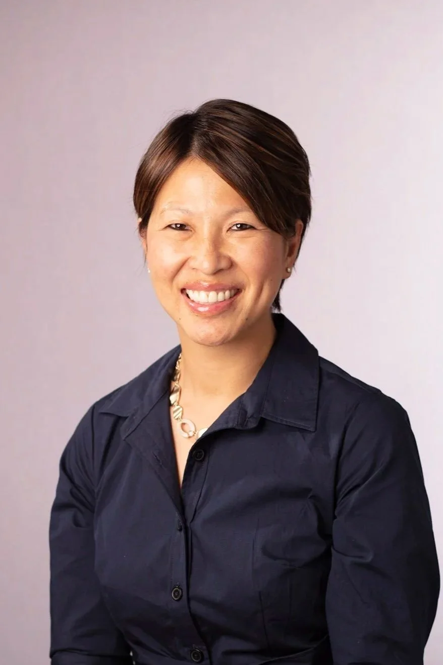 A woman with short, dark hair wearing a black button-up shirt and a necklace, smiling at the camera against a light background.
