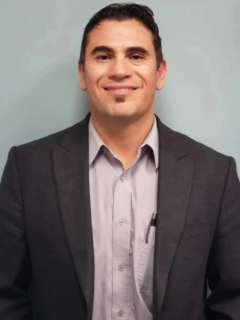 A man smiling in a gray blazer and light gray shirt with a pen in the pocket, standing against a plain wall.