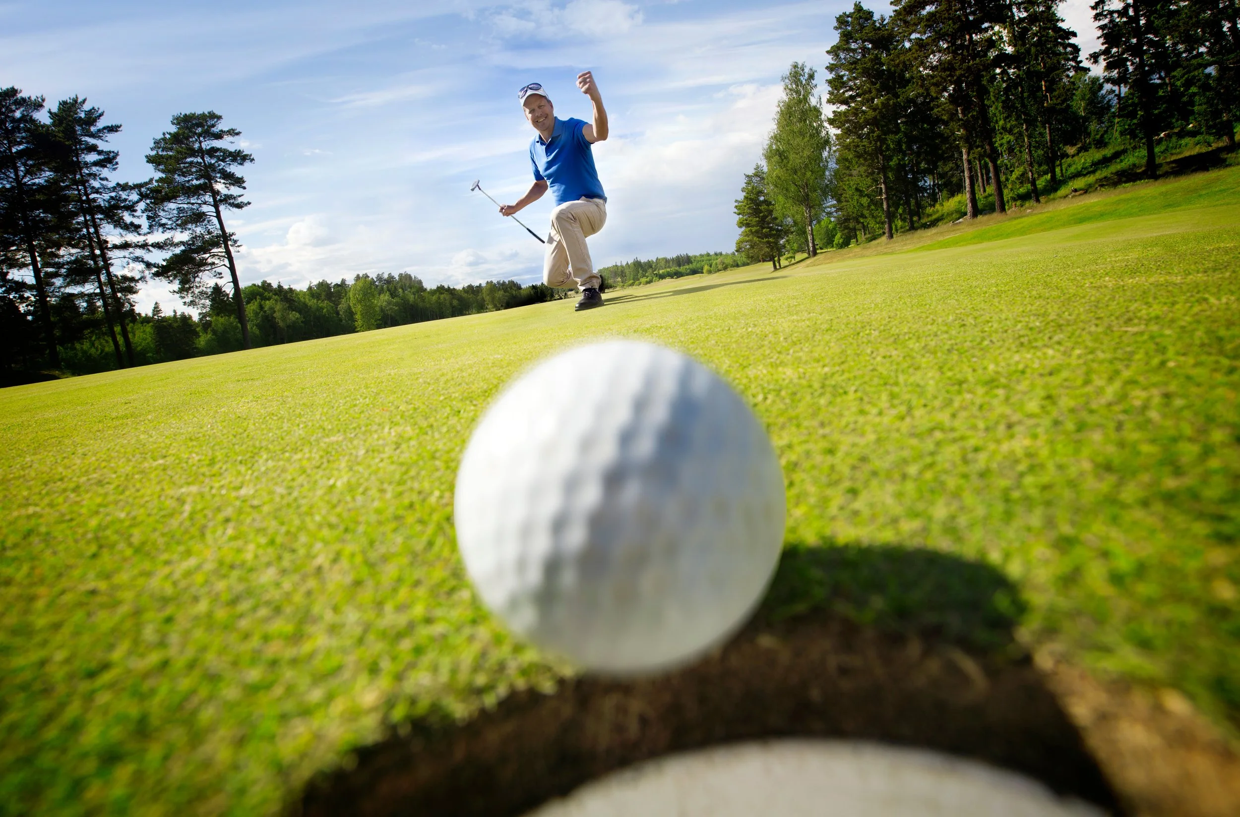 A man in a blue shirt and khaki pants playing golf on a lush green golf course, celebrating after a successful shot, with trees and a bright sky in the background.