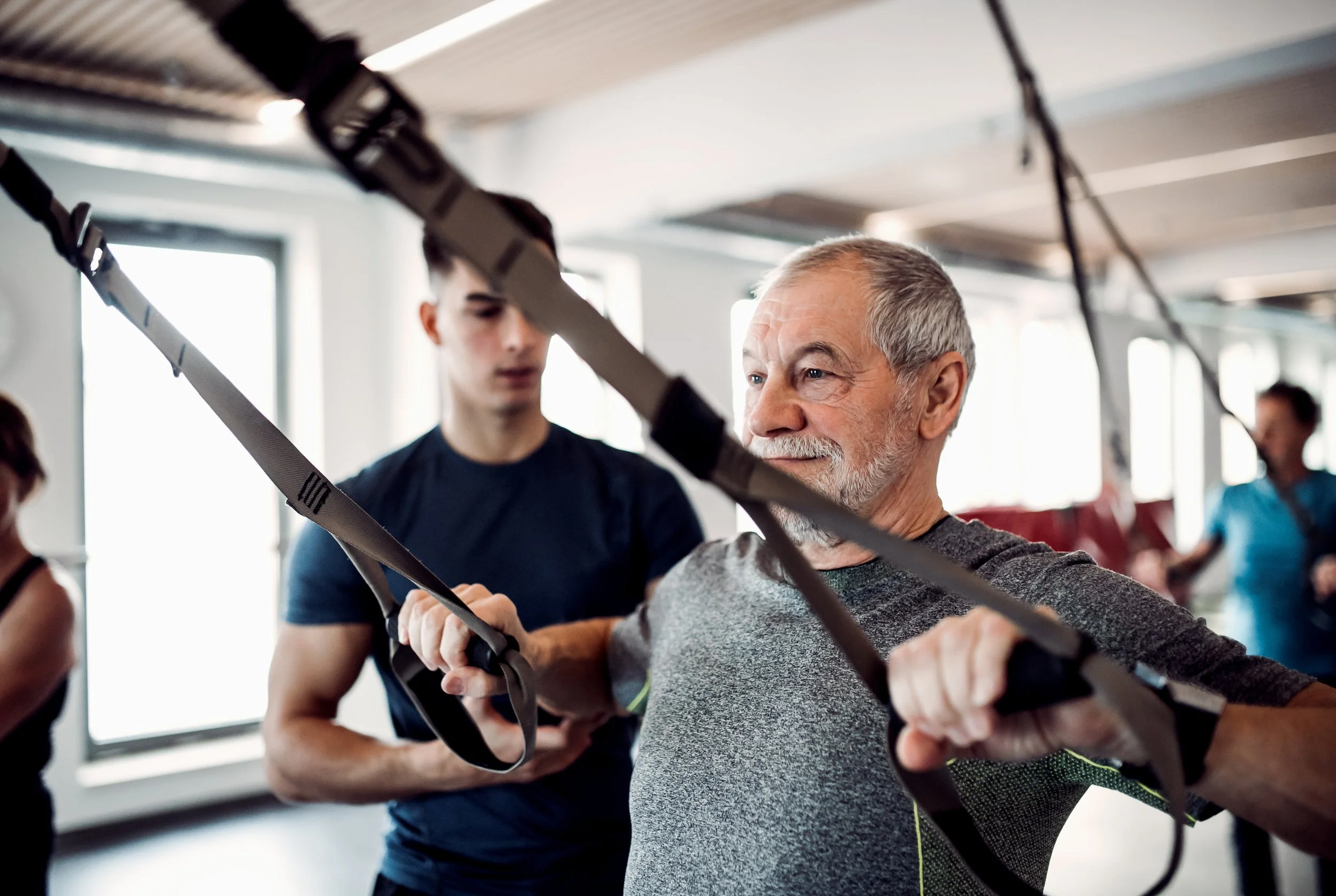 An older man with gray hair and a beard is being assisted by a younger woman with brown hair in a gym or fitness studio. They are practicing an exercise involving straps, possibly suspension training, with windows in the background.