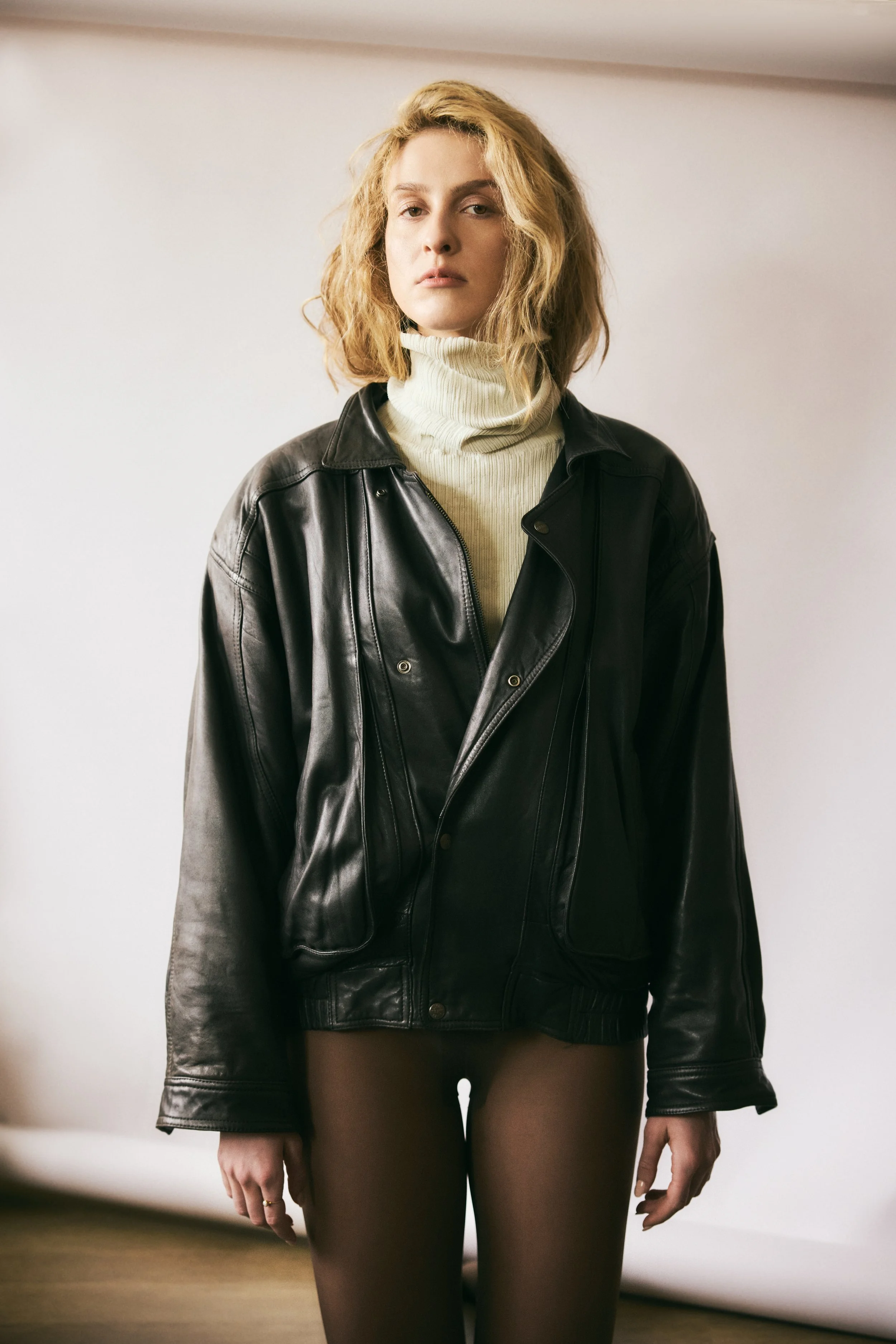 A woman with wavy blonde hair wearing a black leather jacket over a cream turtleneck, standing indoors in front of a plain white wall.