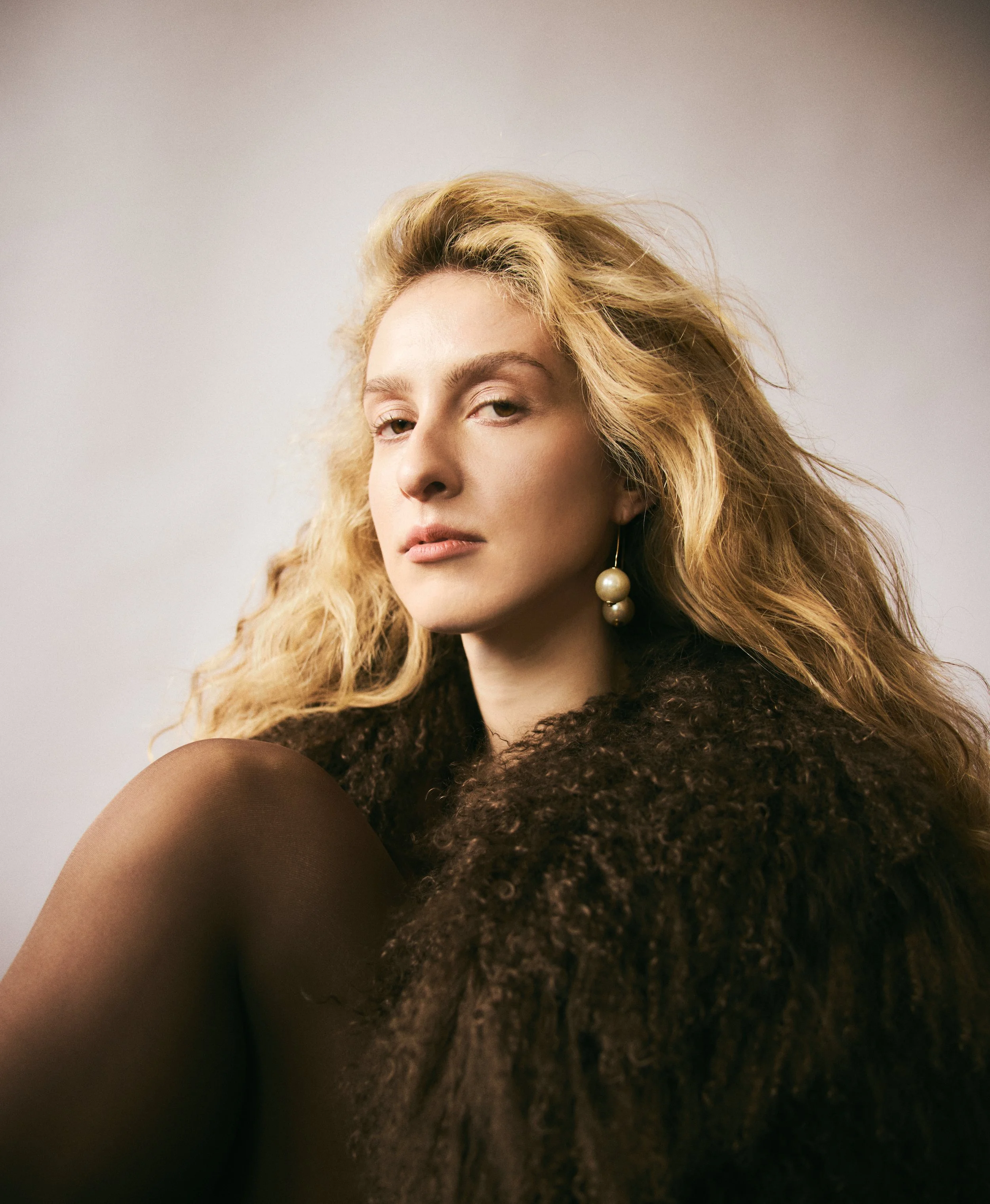 A woman with long blonde curly hair, wearing an earring with multiple pearls and a dark fuzzy sweater, looking confidently at the camera against a plain light background.