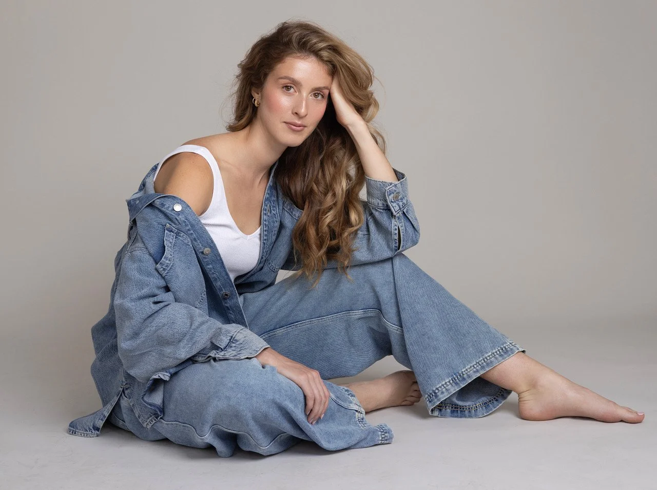 A woman with long wavy hair sitting on the floor, wearing a denim jacket and matching jeans, with a white tank top. She is barefoot, with one hand resting on her knee and the other supporting her head, against a plain light gray background.