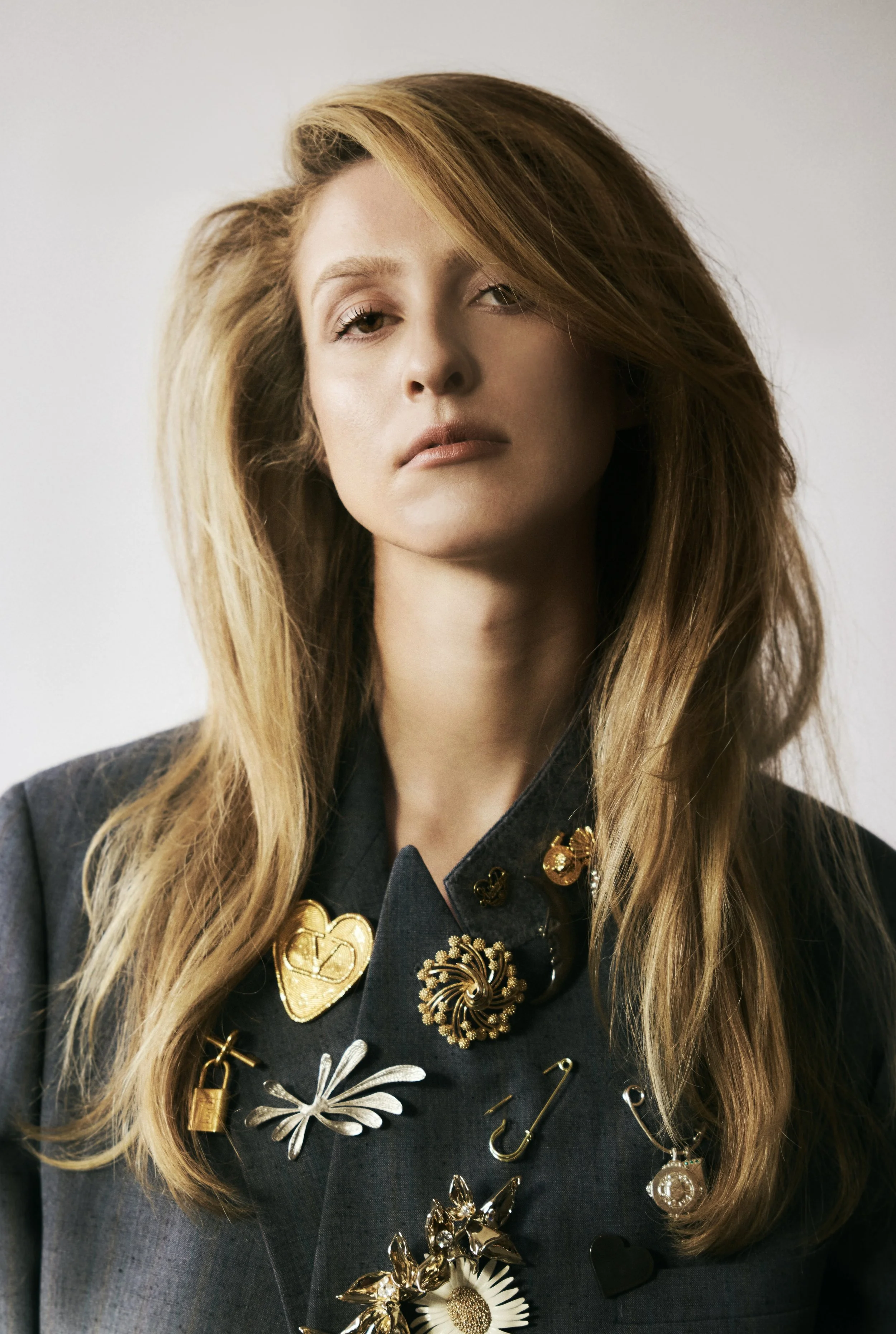 A woman with long, light brown hair wearing a gray jacket adorned with various metallic and decorative pins and brooches in an editorial portrait.
