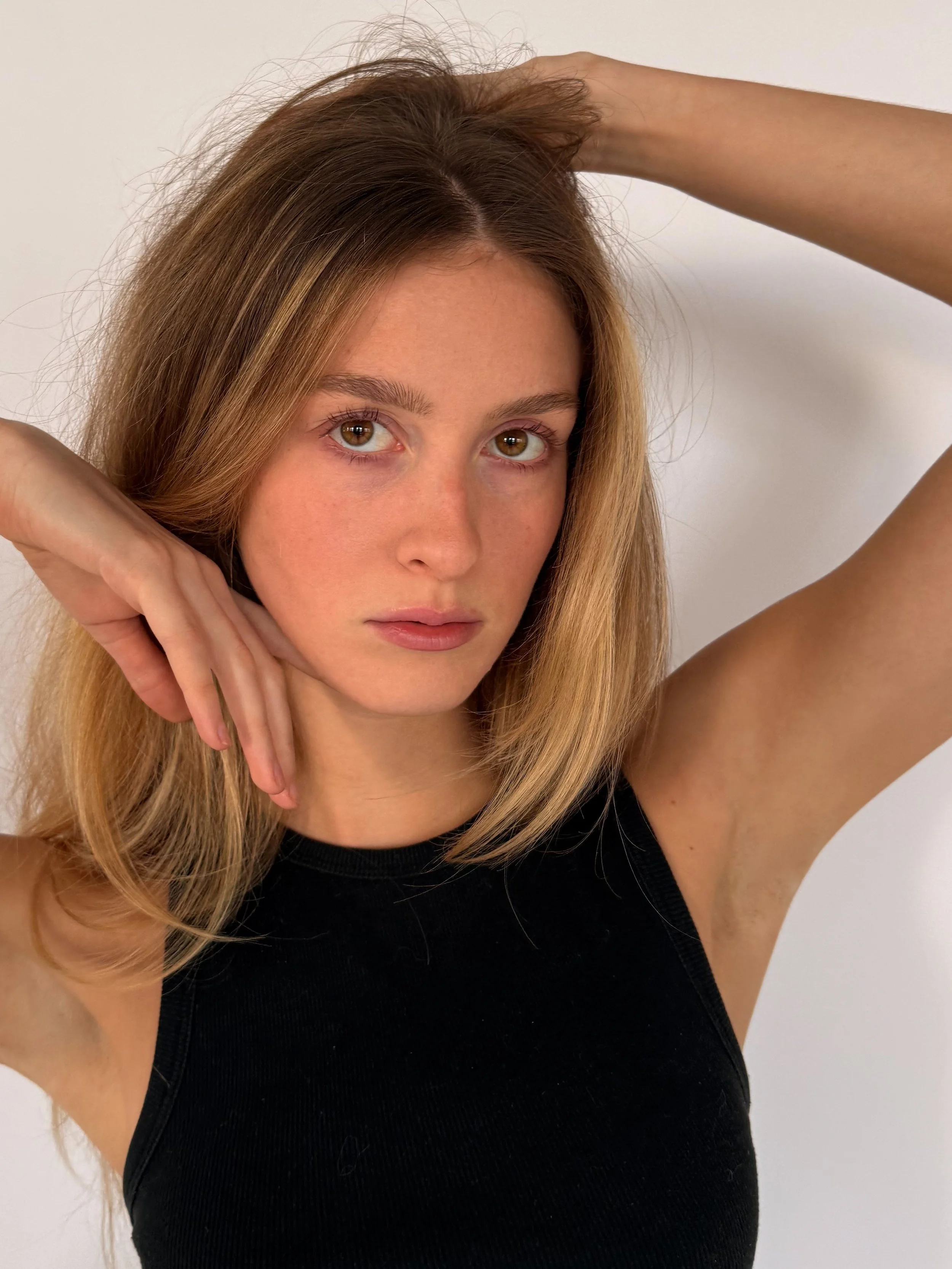 A woman with light brown hair in a black sleeveless top poses with her arms raised, one hand resting on her head and the other near her face, against a white background.