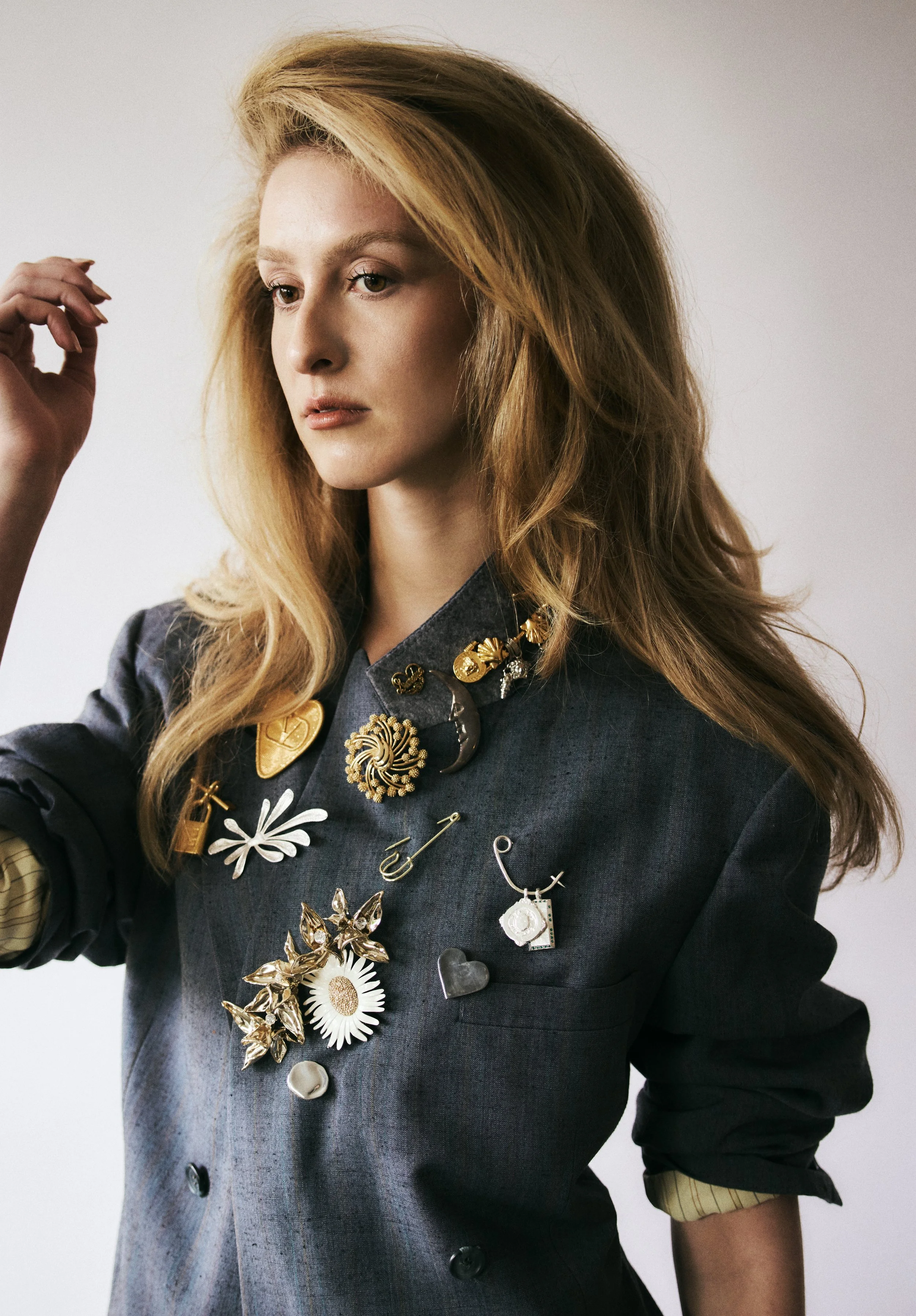 A woman with long, wavy blonde hair wearing a dark gray blazer decorated with various jewelry pins, including flowers, hearts, and abstract shapes, against a plain background.