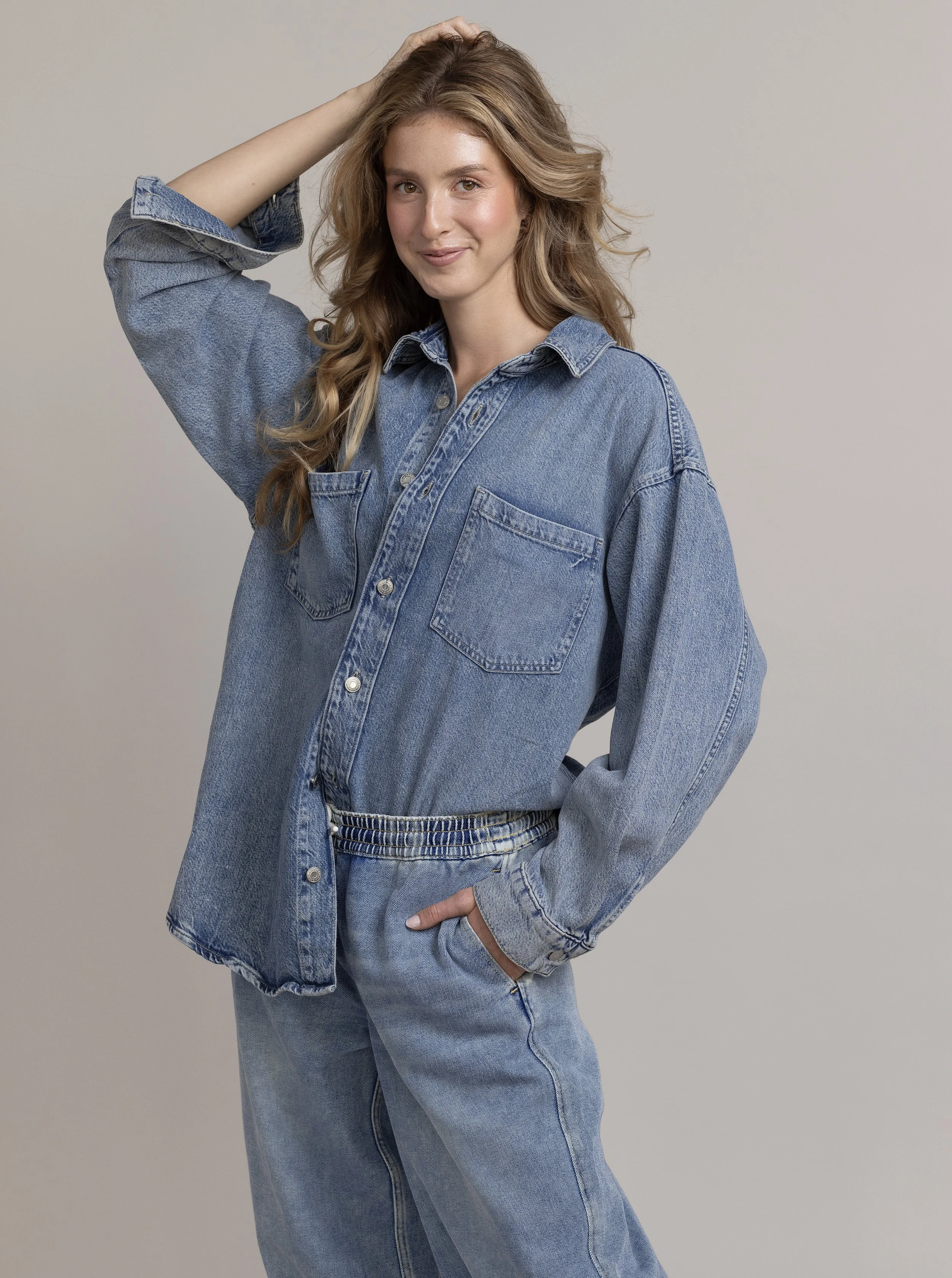 A young woman with long, wavy, light brown hair wearing a oversized denim shirt and matching jeans, smiling, with her left hand in her pocket and her right hand touching her head, standing against a plain gray background.