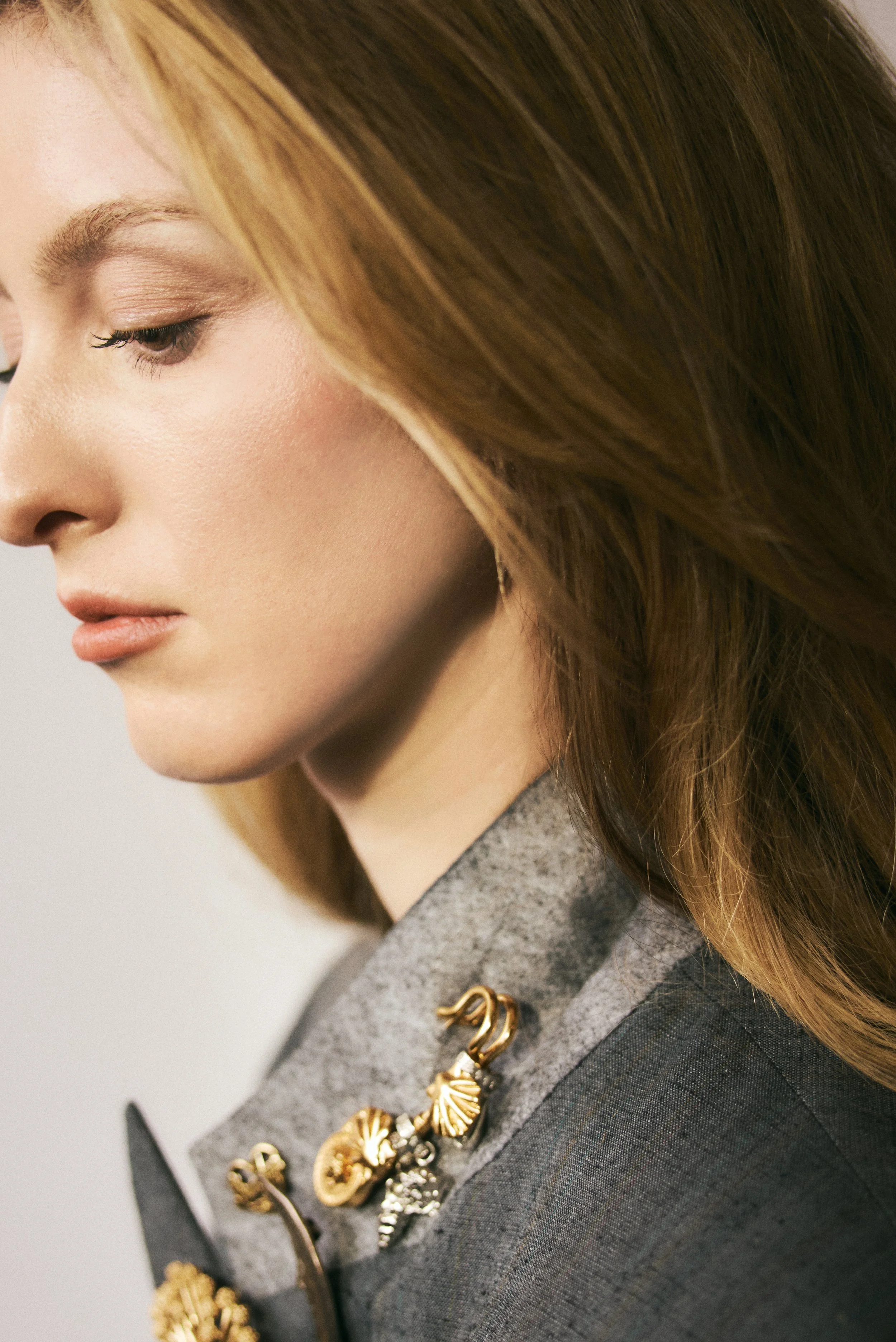 Close-up of a woman with light skin, red hair, and makeup, wearing a gray blazer with decorative gold and silver pins.
