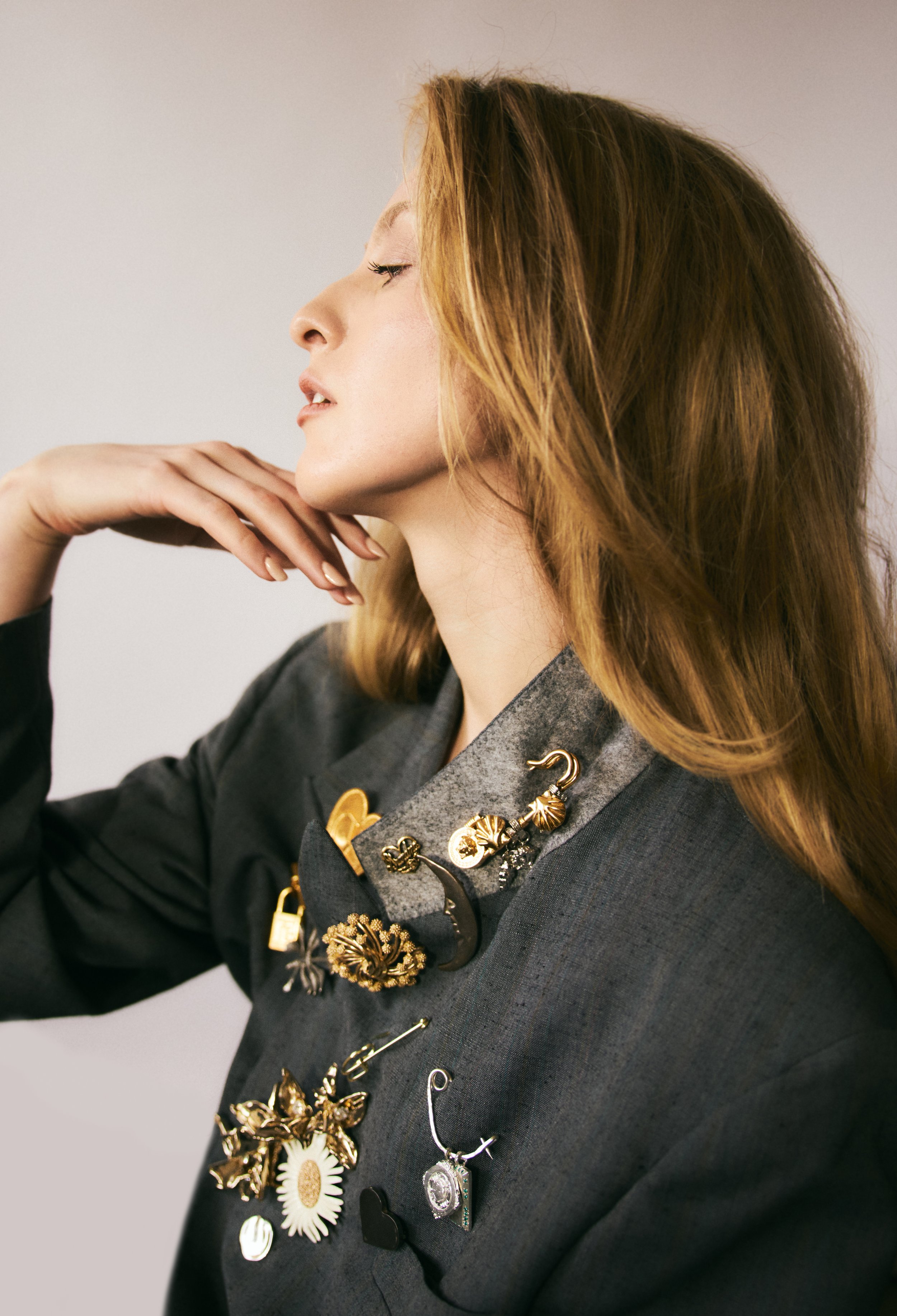 A woman with red hair in profile, wearing a dark gray blazer decorated with various gold, silver, and black jewelry pins and brooches.