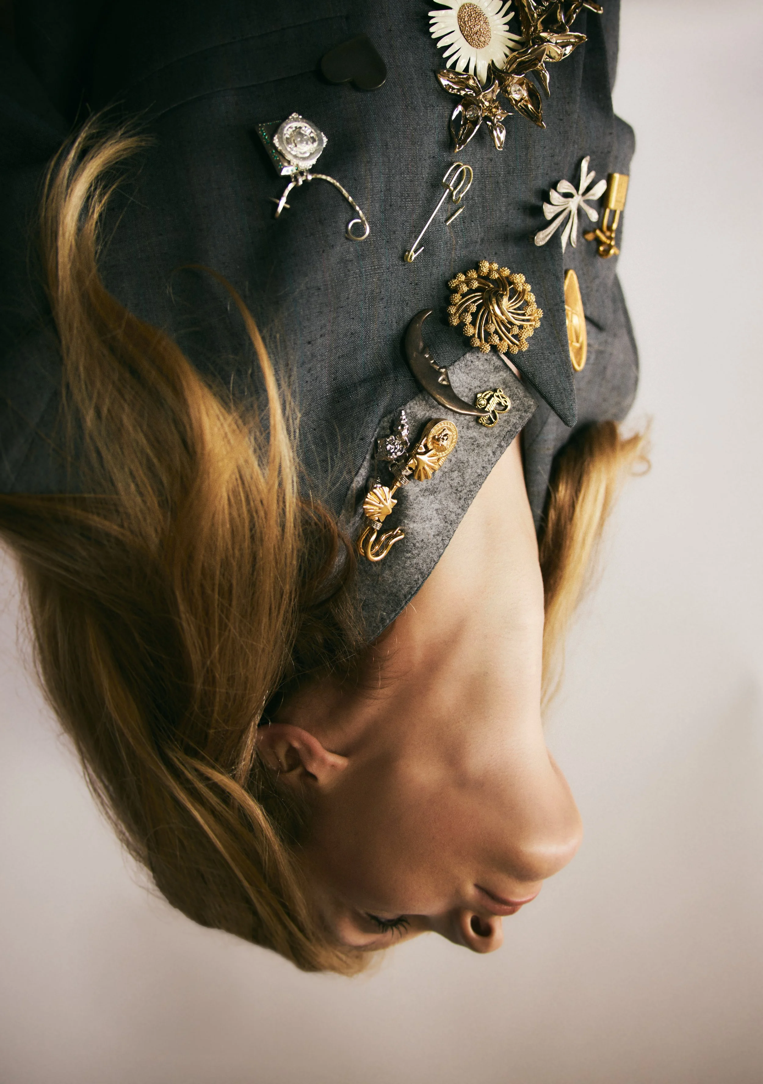 A woman with long red hair and light skin, dressed in a dark blazer adorned with various decorative pins and brooches, is positioned sideways with her head tilted downward.
