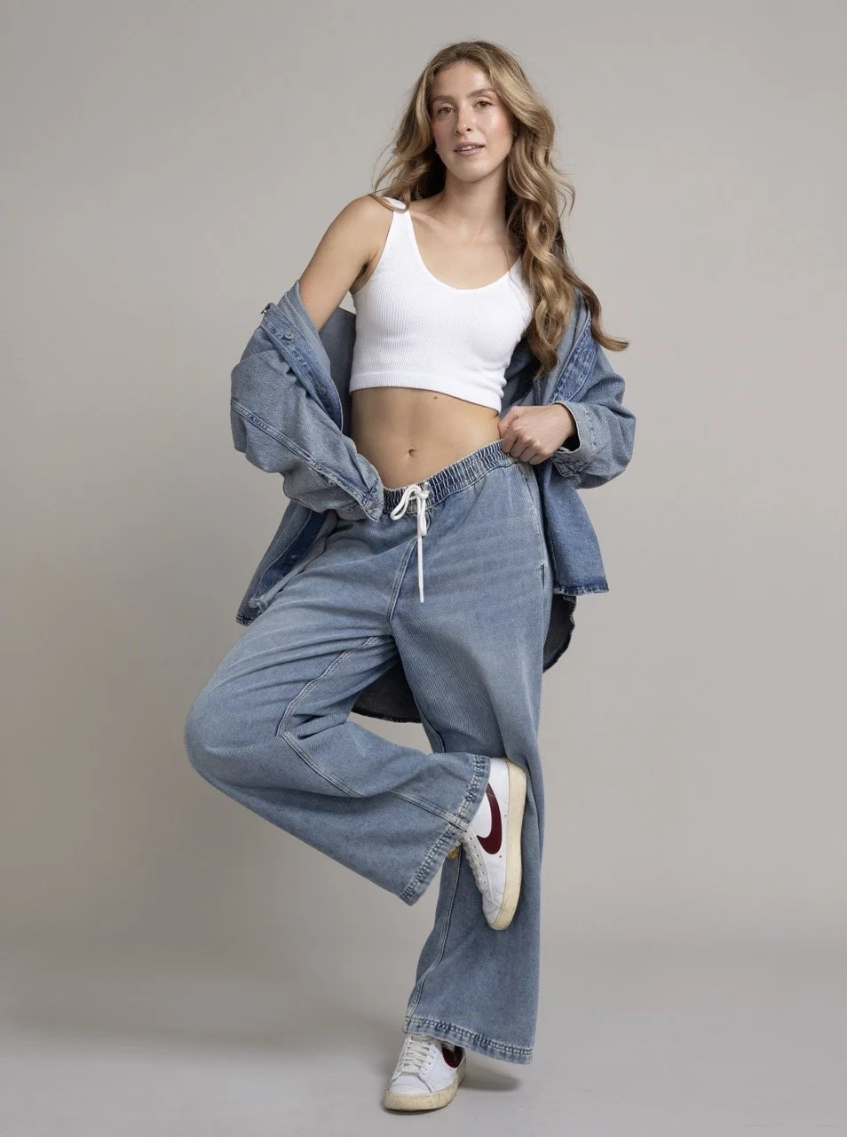 A young woman with long wavy blonde hair poses against a plain gray background. She is wearing a white crop top, loose-fitting blue jeans with a drawstring, white sneakers with red accents, and a blue denim jacket draped over her shoulders. She has o