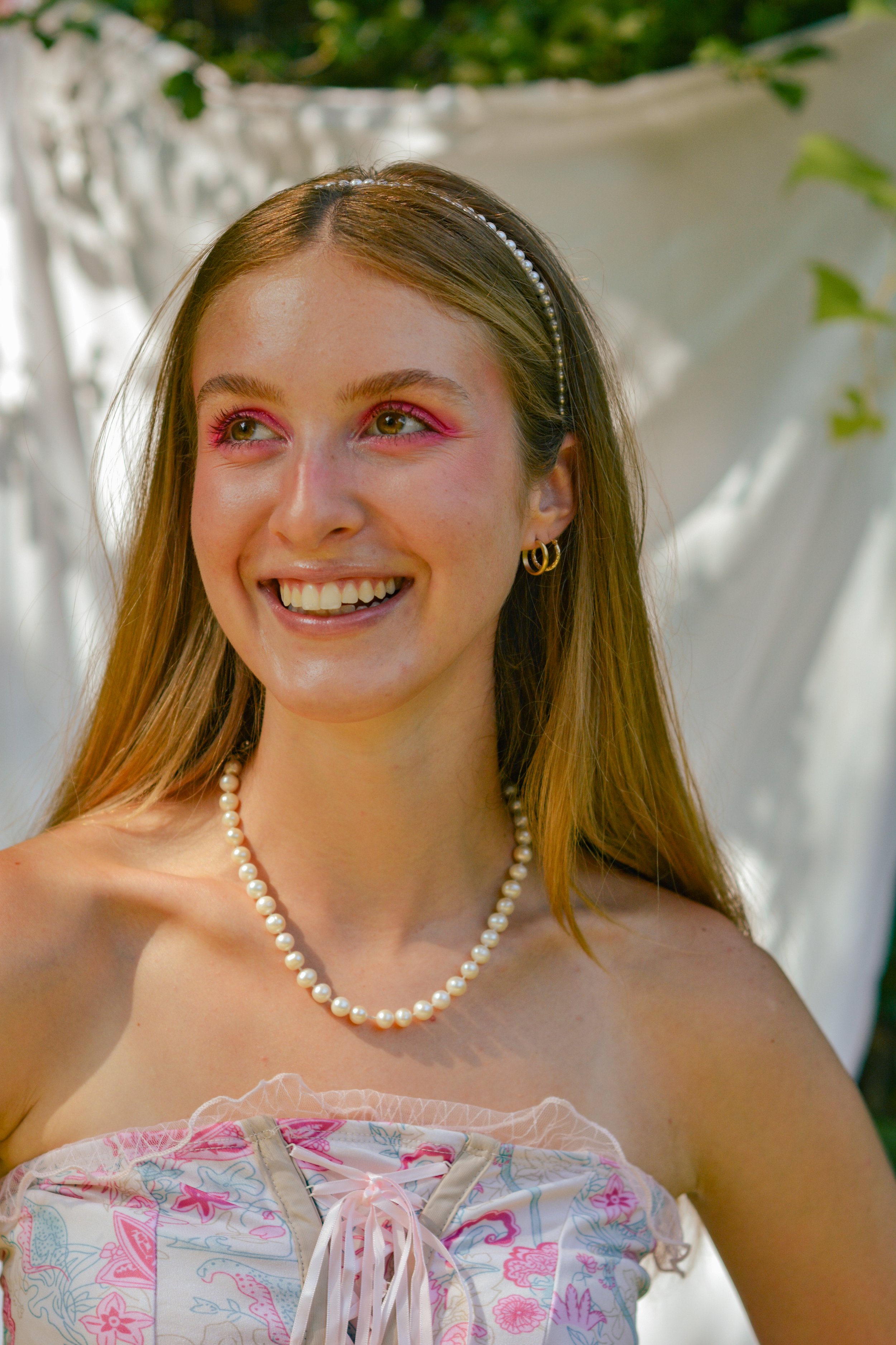 A young woman smiling outdoors, wearing a string of pearls, gold hoop earrings, a pearl headband, and a pink floral strapless dress with pink makeup.
