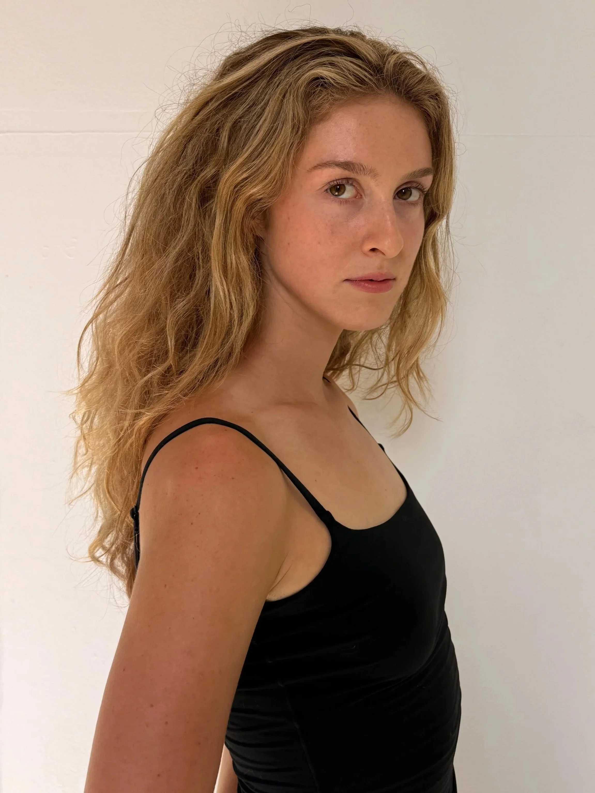 A young woman with wavy red hair wearing a black spaghetti strap top standing against a plain white background.