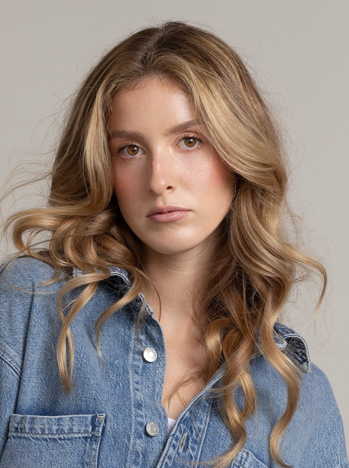 A young woman with long, wavy, light brown hair wearing a denim jacket against a neutral background.